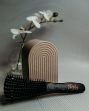 Load image into Gallery viewer, Black hairbrush with 'Hairflax' branding on a textured surface with a beige towel and white flowers in the background.