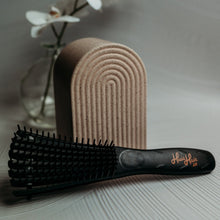 Load image into Gallery viewer, A pink and black Huia hair detangling brush with curved comb teeth, placed on a white surface next to a cake stand and a vase with flowers.
