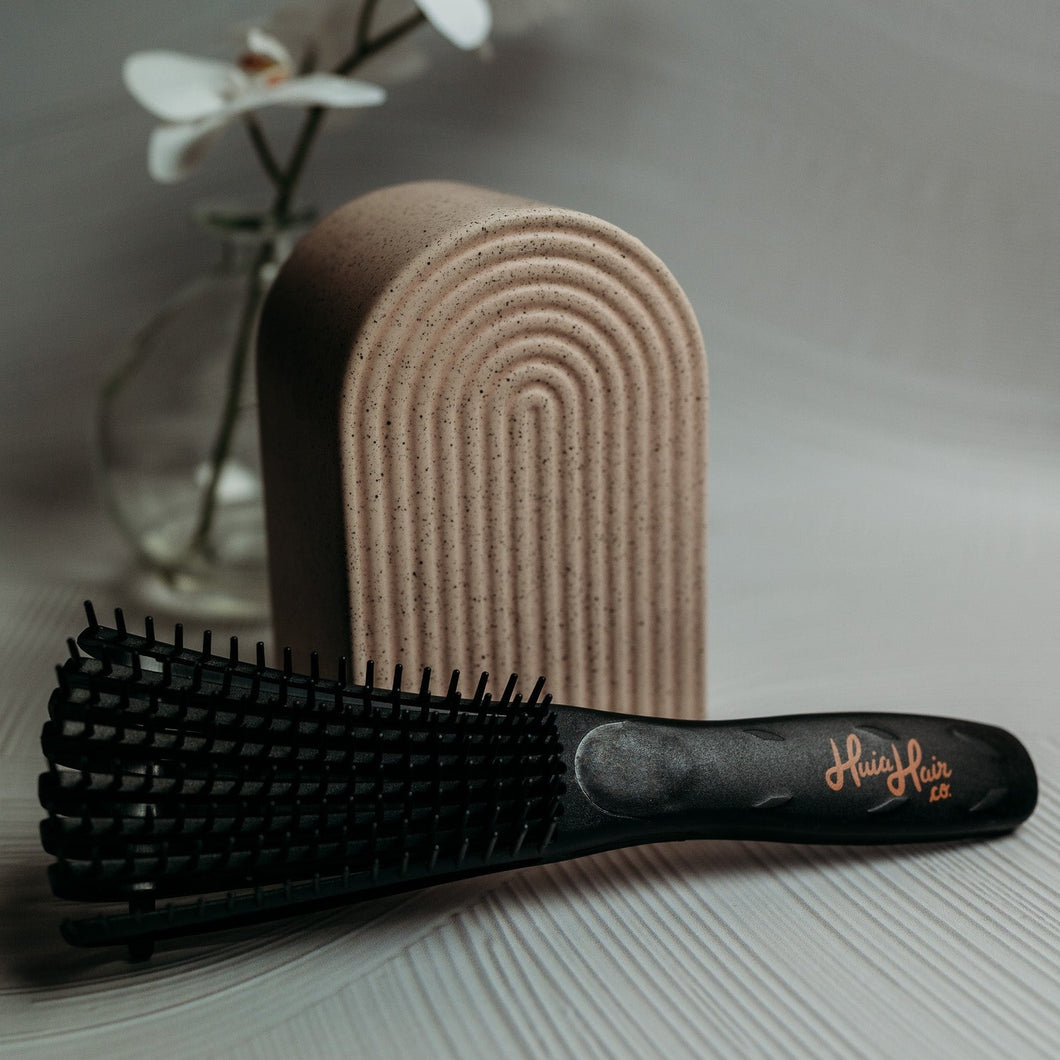 A pink and black Huia hair detangling brush with curved comb teeth, placed on a white surface next to a cake stand and a vase with flowers.