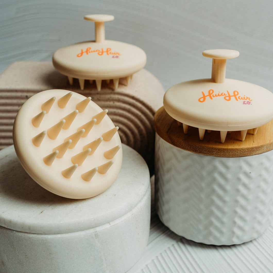 A silicone scalp massage brush with a beige handle and bristles, placed on a textured surface.